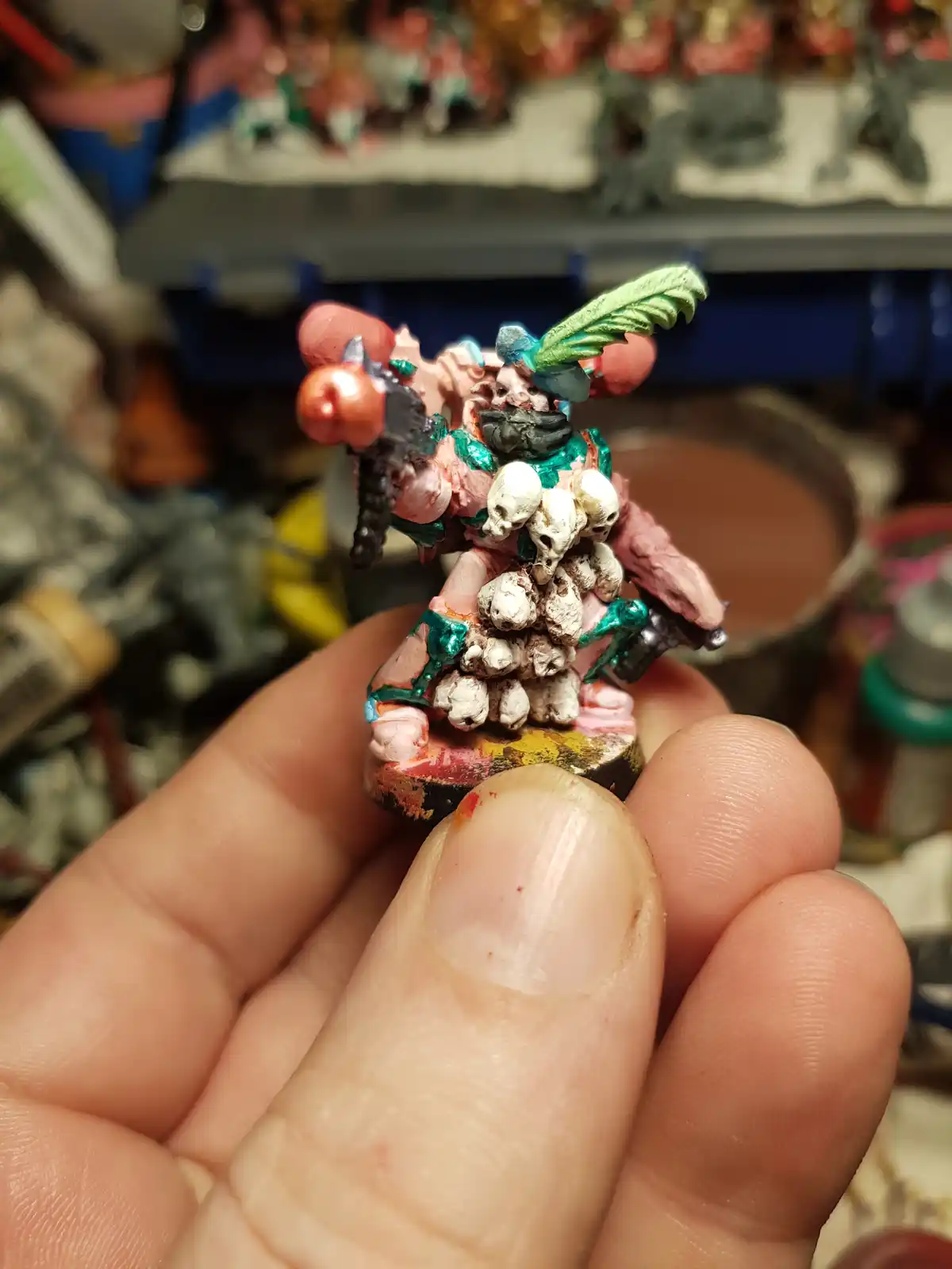 A hand holds a poorly painted miniature figurine, which appears grotesque and unfinished, covered in pink, green, and white…