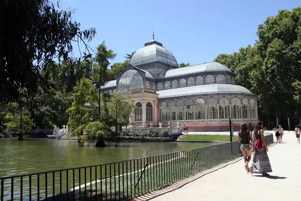 A sunny photo of the Crystal Palace in Madrid's Retiro Park, a large ornate glass and iron greenhouse surrounded by trees…