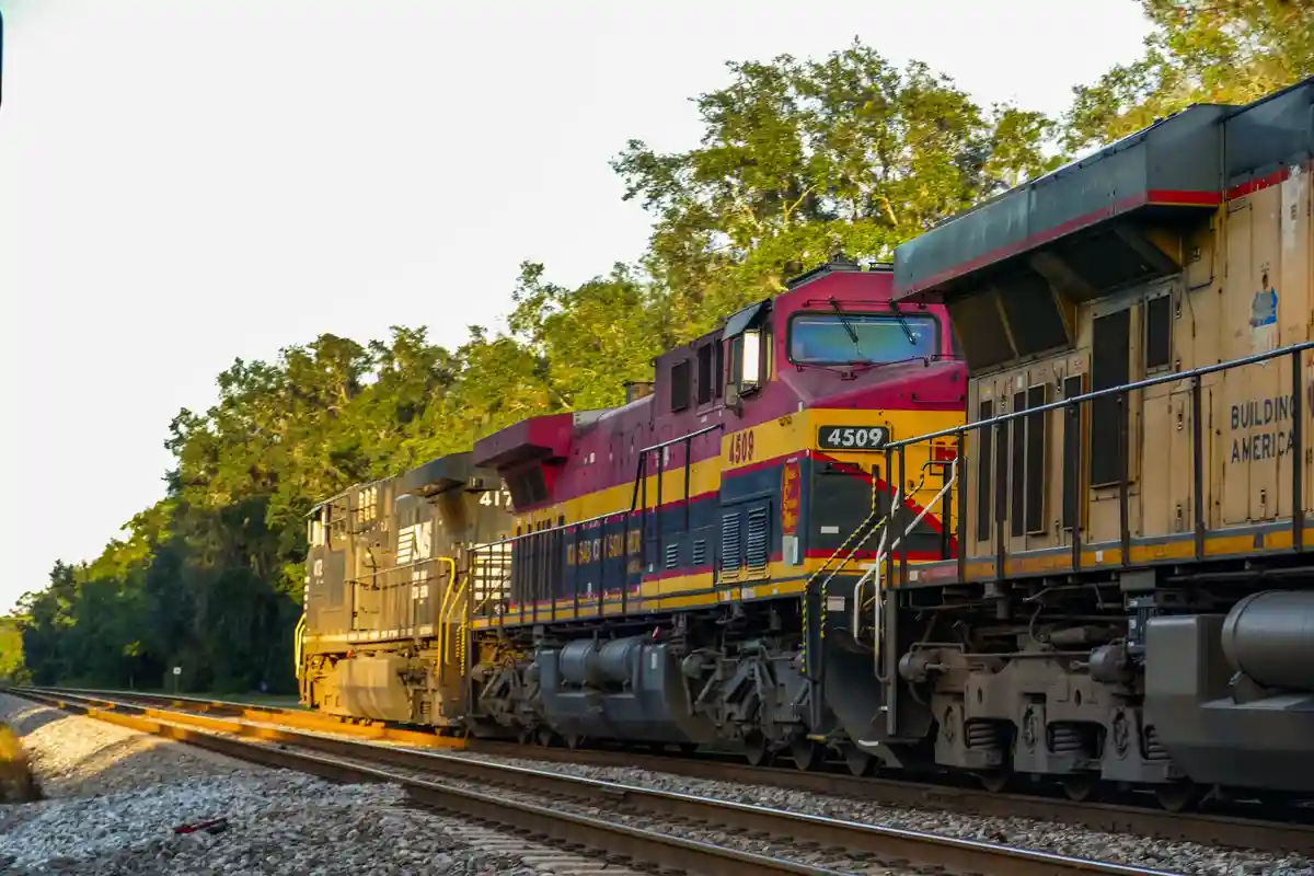 A Kansas City Southern freight train with locomotives numbered 4509 and 417 travels along tracks through a wooded area. The…