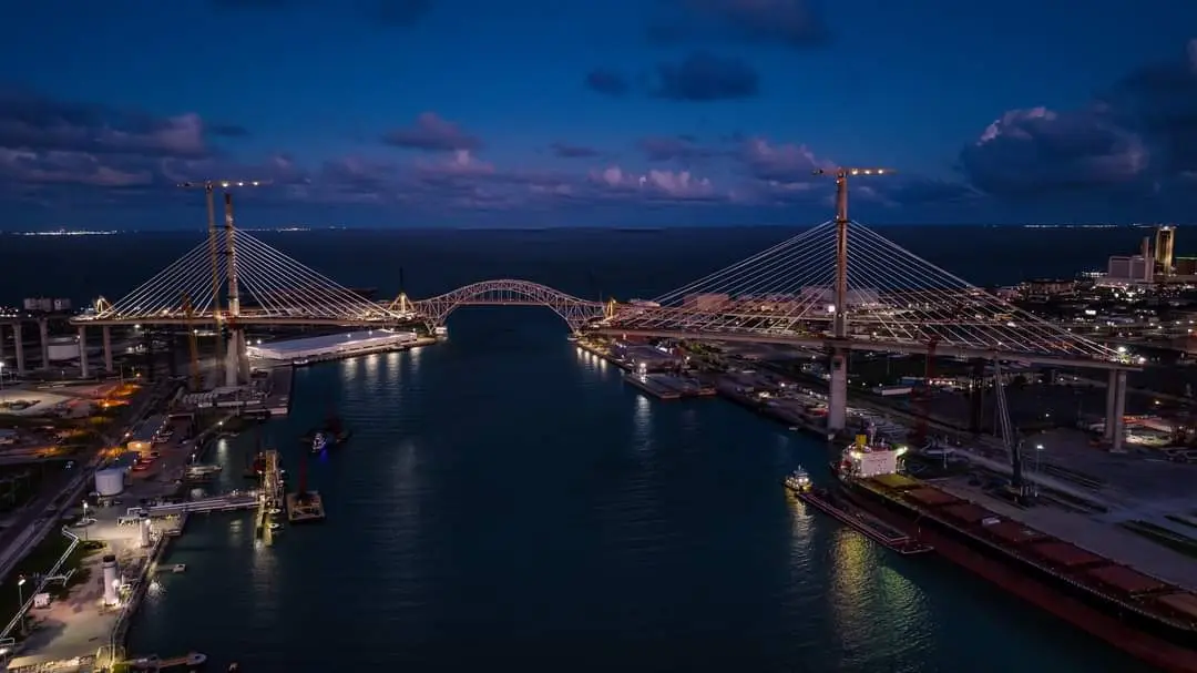 A nighttime aerial photo of a large, illuminated cable-stayed bridge spanning a waterway, with industrial port areas and…