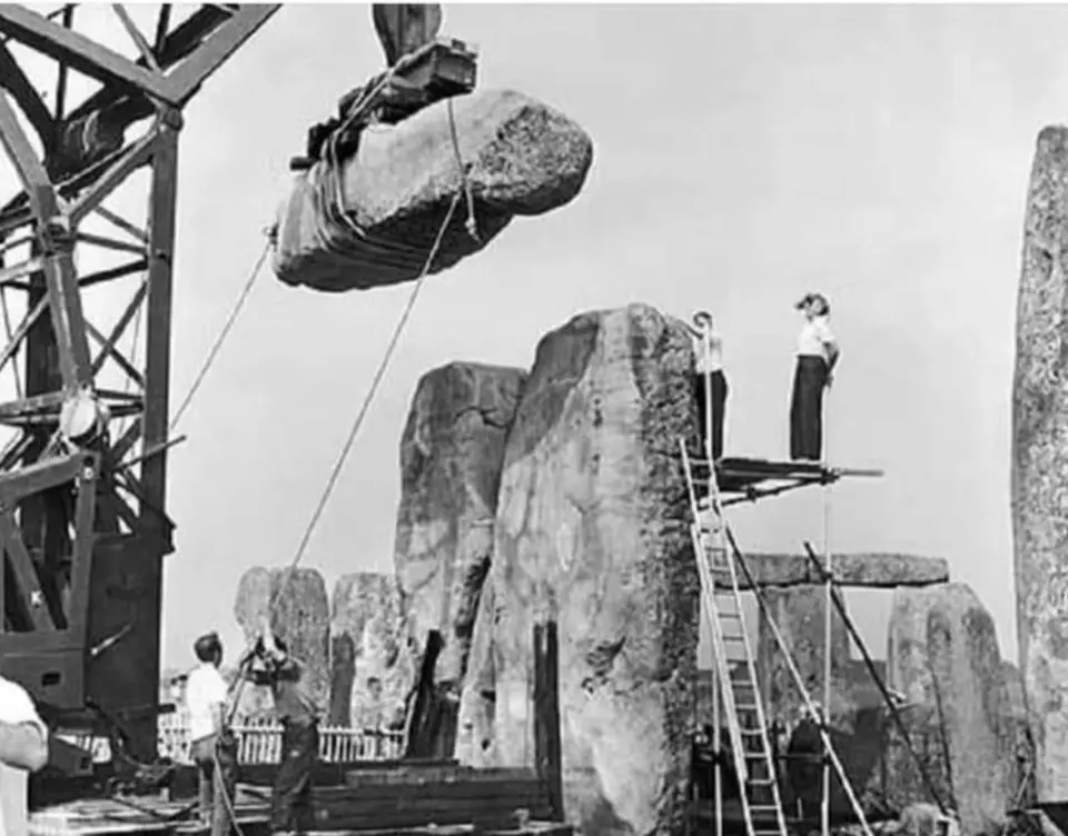A black-and-white historical photo shows workers using a crane to lift a massive stone block near other standing stones…