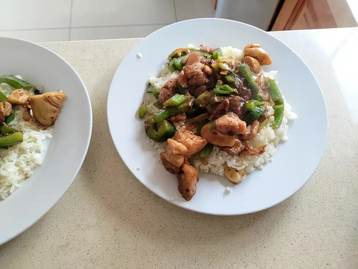 Two white plates sit on a light-colored countertop, each holding a serving of chicken and green pepper stir-fry over white…