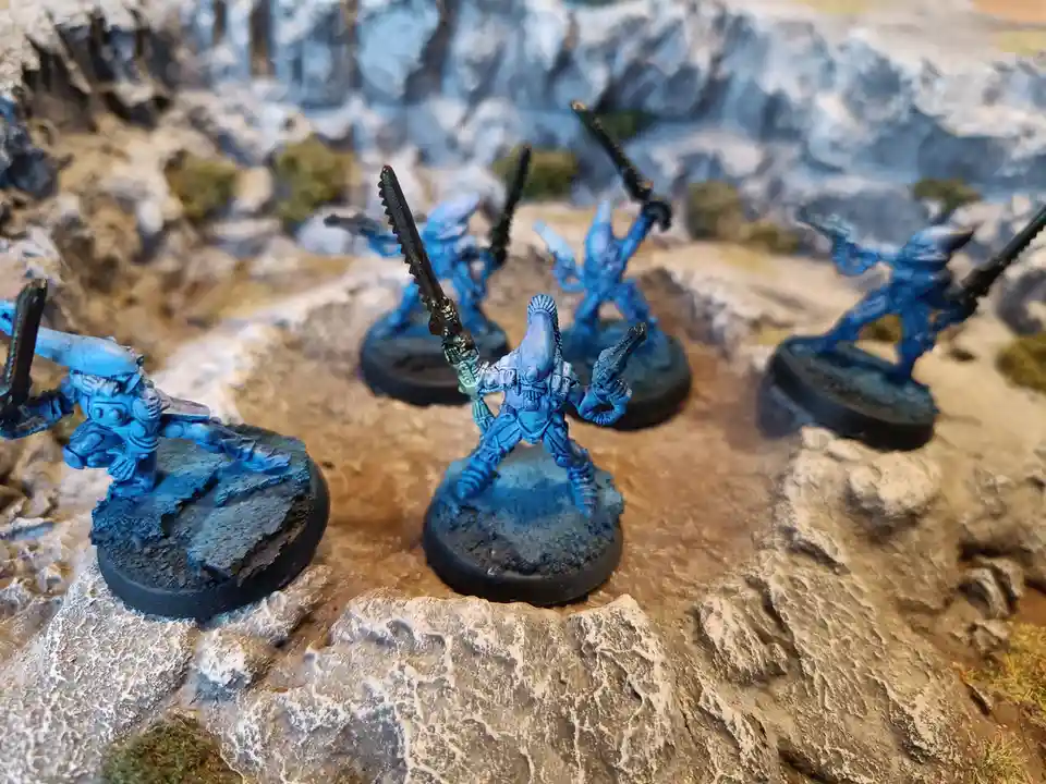 Several blue-painted miniature alien warriors with weapons stand on rocky terrain. The central figure holds a large serrated…