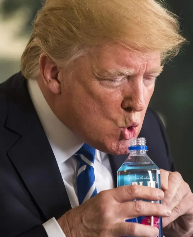 A close-up photo shows a man with blond hair, wearing a dark suit and blue tie, drinking from a clear plastic water bottle…