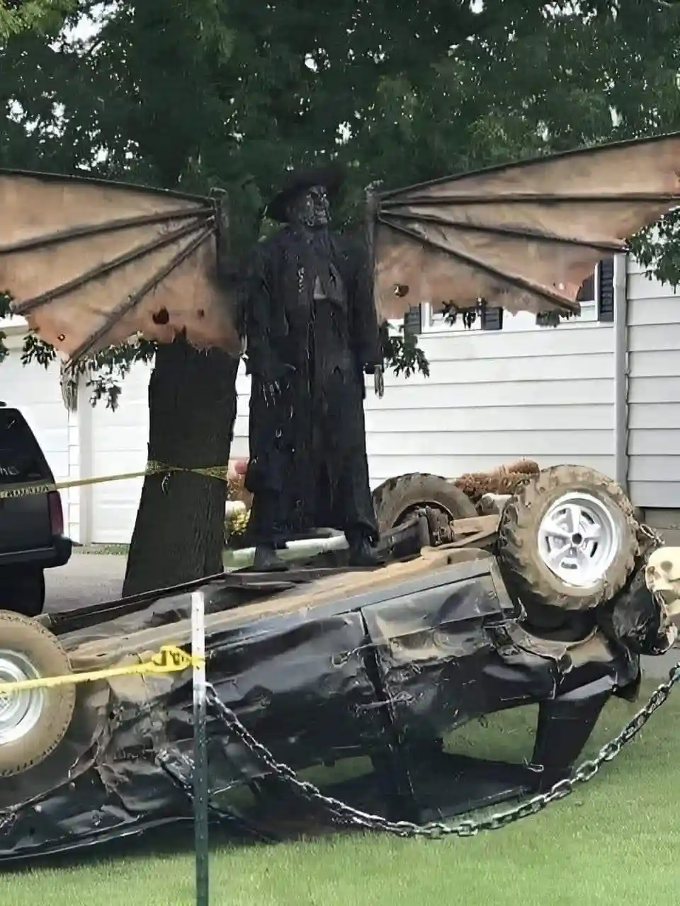 a large, dark, winged dragon statue stands atop a flipped black car, which is surrounded by yellow caution tape and a chain…