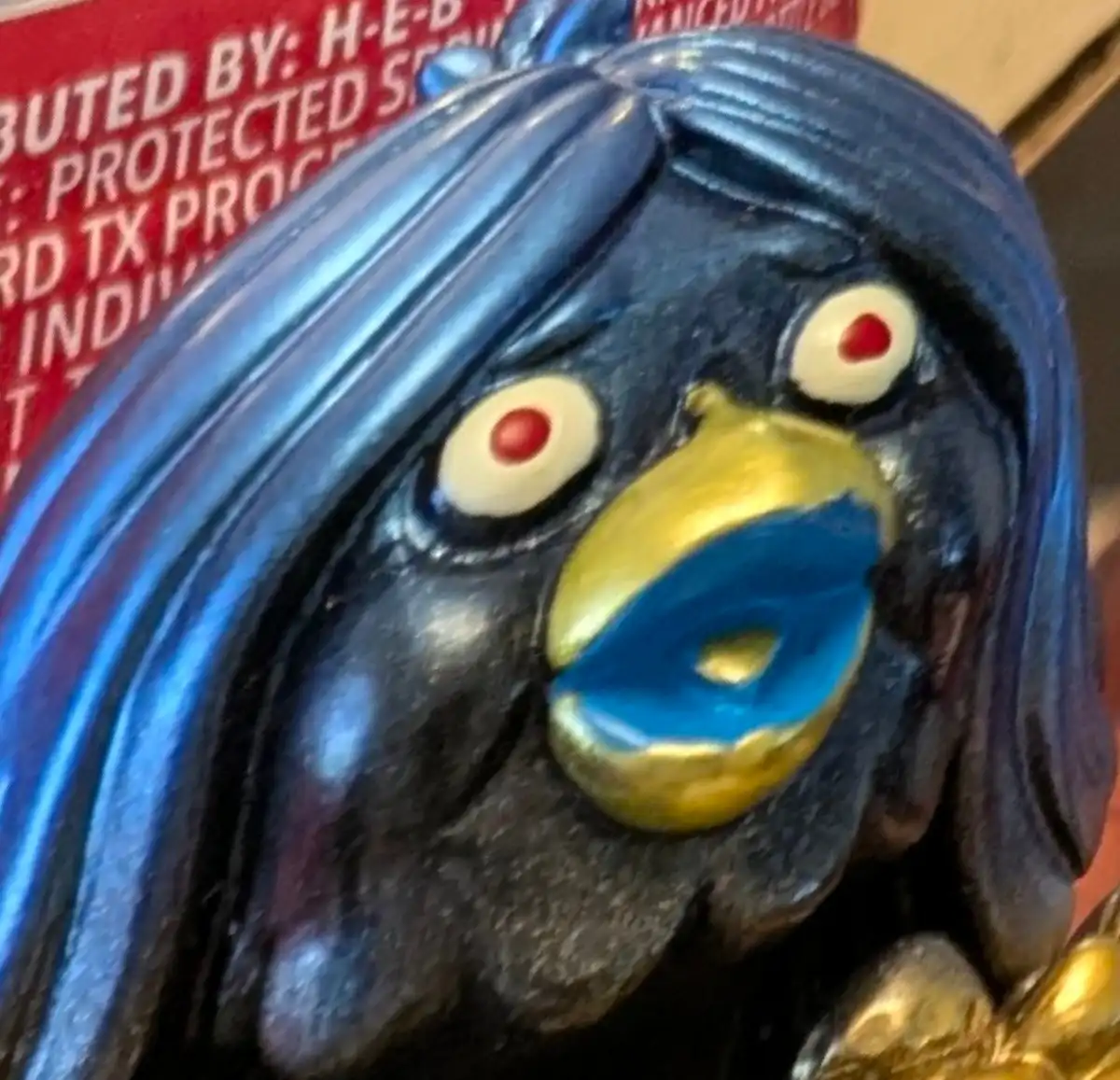 A close-up of a black plastic figurine resembling a bird with blue hair, red dot eyes, and a gold beak holding a blue donut…