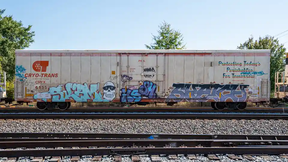 A weathered Cryo-Trans refrigerated railcar, covered in graffiti including a skull wearing sunglasses, sits on tracks under…