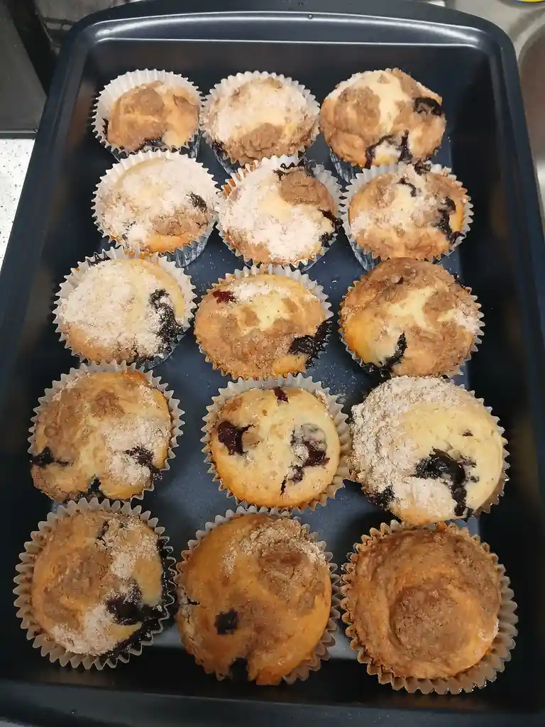 A black baking tray holds 15 freshly baked blueberry muffins in white paper liners, some topped with sugar and cinnamon…