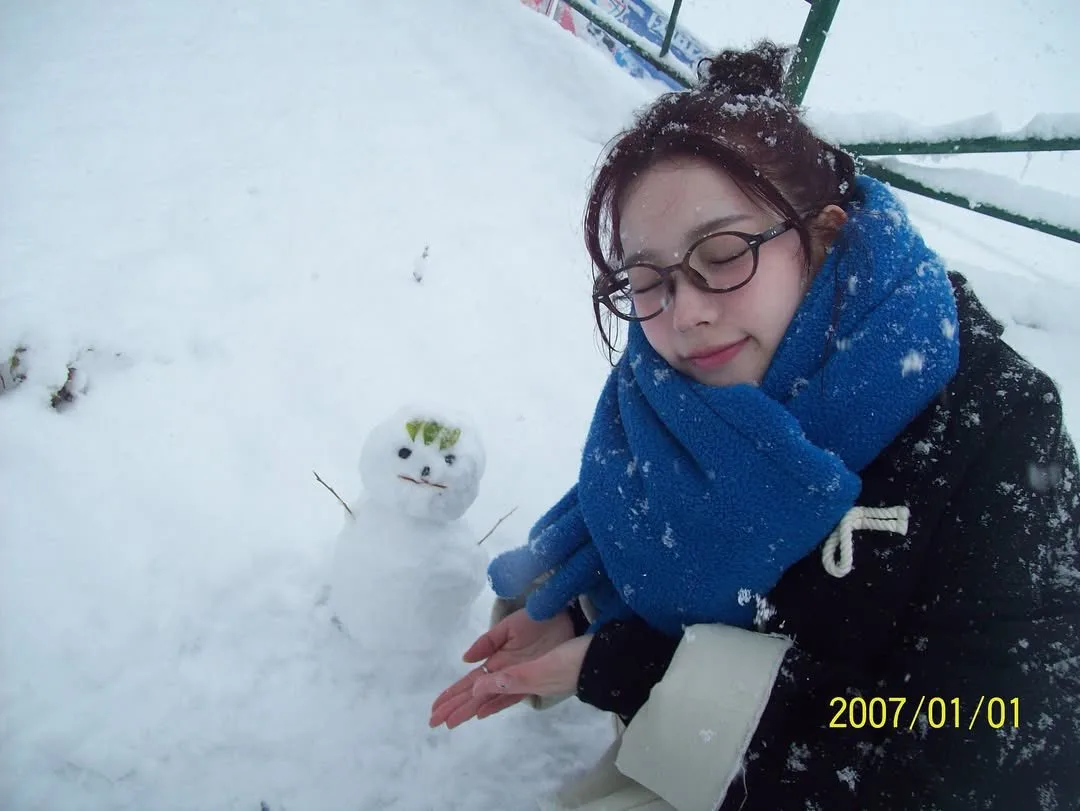 A young woman with glasses and a blue scarf sits in deep snow, smiling gently with eyes closed as she extends her hands…