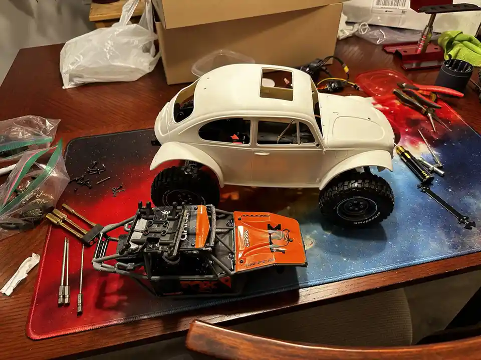 A white remote-controlled Volkswagen Beetle model is shown on a red and blue galaxy-patterned mousepad, partially assembled…
