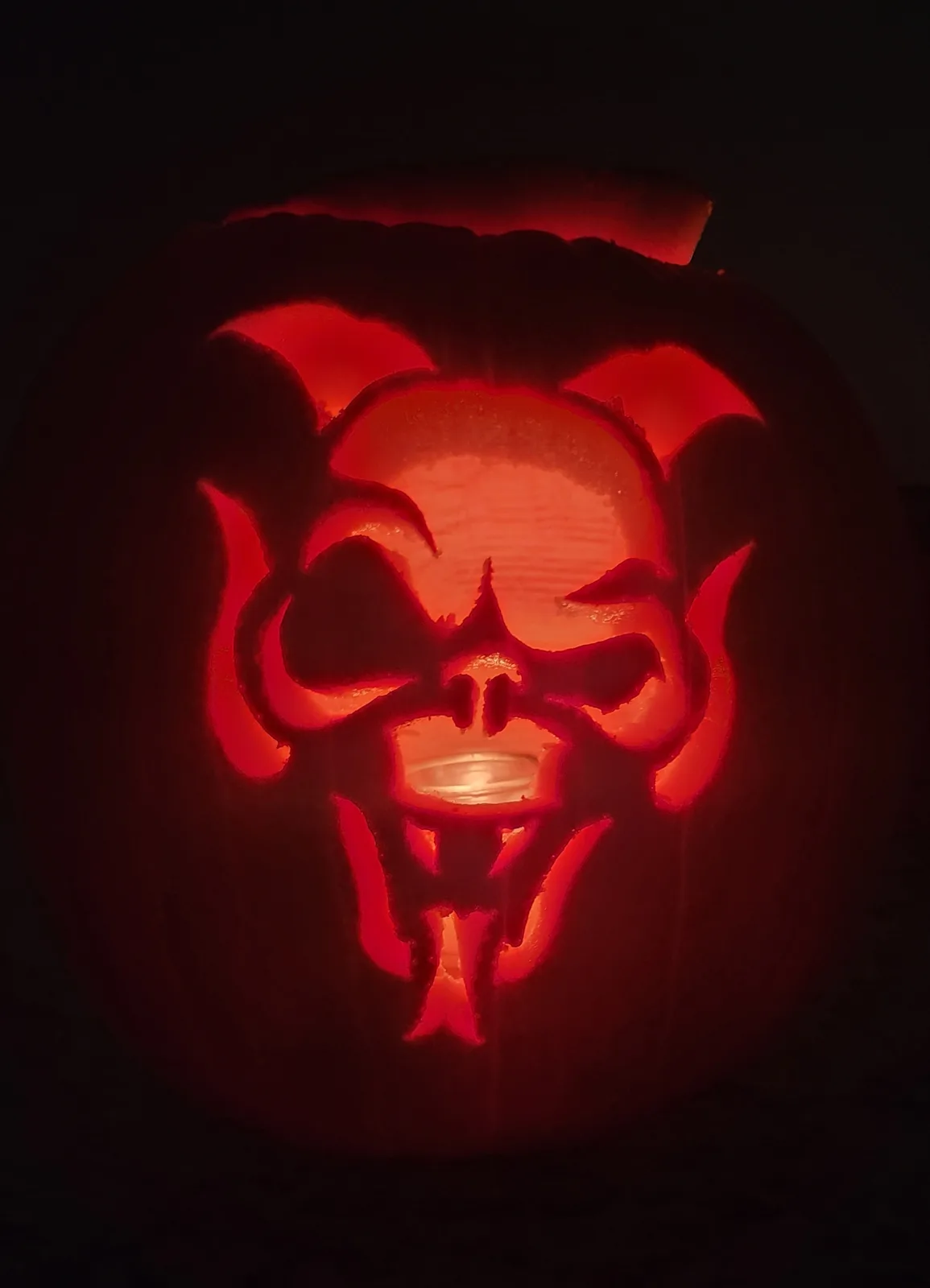 A carved pumpkin illuminated from within, featuring a demonic face with horns, sharp teeth, and a sinister grin, glowing red…