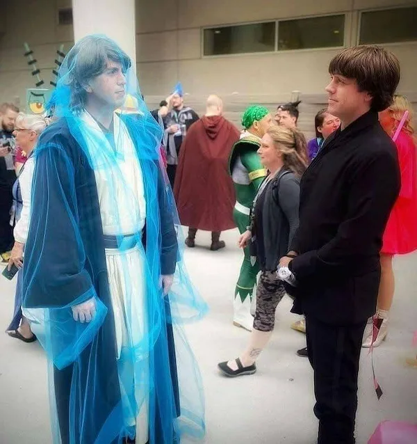 Two men dressed as Star Wars characters stand facing each other in a crowded convention hall. The man on the left wears a…