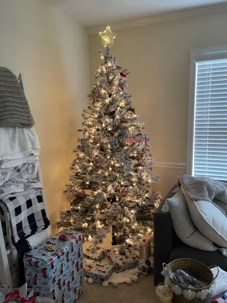 A flocked Christmas tree decorated with warm white lights, pink ornaments, and a star topper stands in a living room corner…