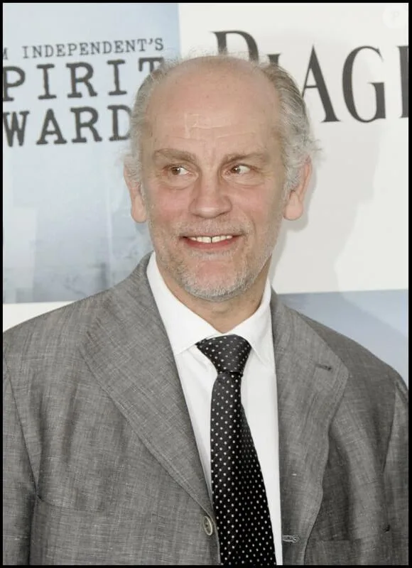 A man with gray hair and a beard, wearing a gray suit and polka-dot tie, smiles while looking sideways at the camera. Behind…