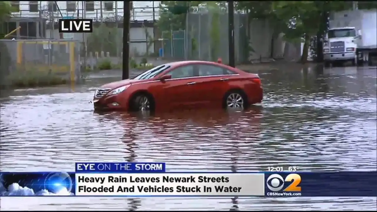 A red sedan is partially submerged in floodwater on a city street, with a news broadcast overlay showing 'LIVE', 'EYE ON THE…
