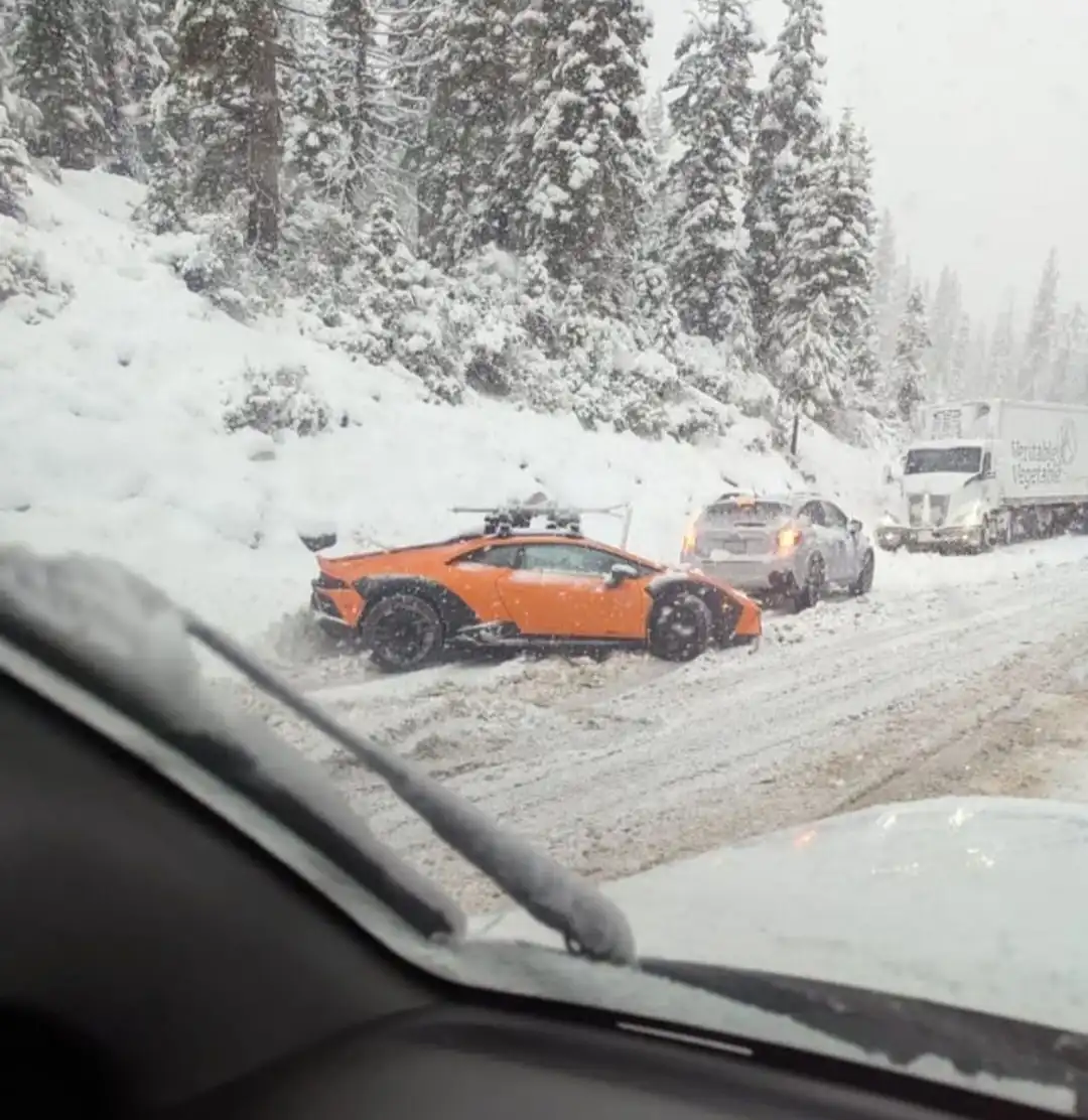 A bright orange Lamborghini sports car is stuck sideways in deep snow on a snowy mountain road, with a silver SUV and a…