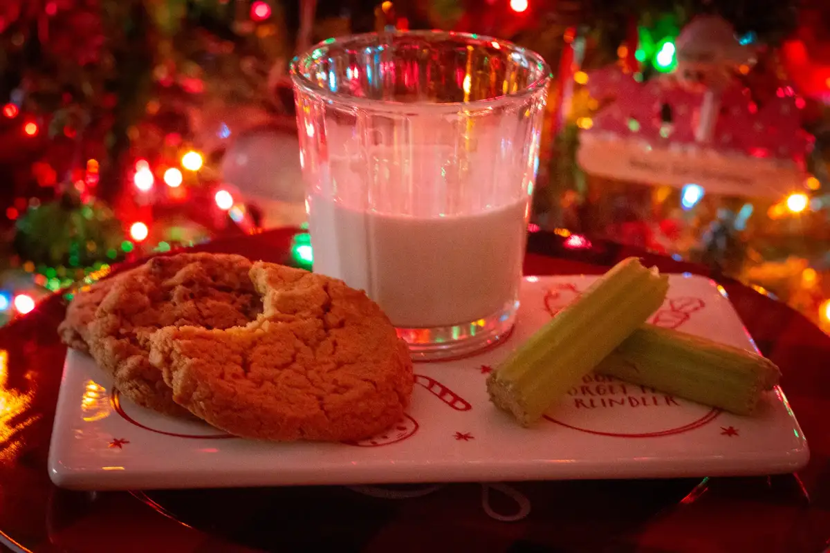 A festive plate holds a glass of milk, a partially eaten cookie, and two celery sticks, set against a blurred background of…