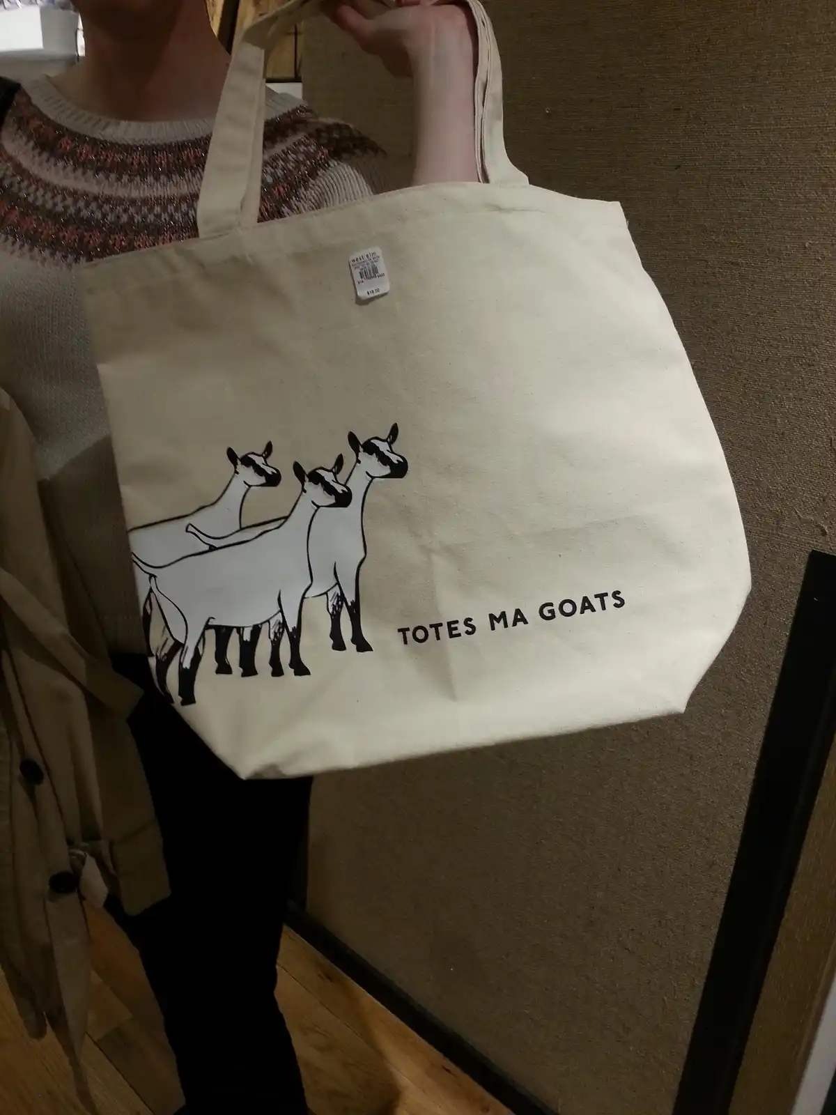 A person holds up a beige canvas tote bag featuring a black-and-white graphic of three goats and the text 'TOTES MA GOATS'…