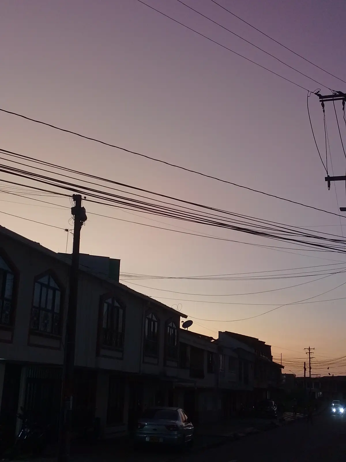 A street scene at dusk with silhouetted houses, power lines, and a car parked on the side. The sky shows a gradient from…