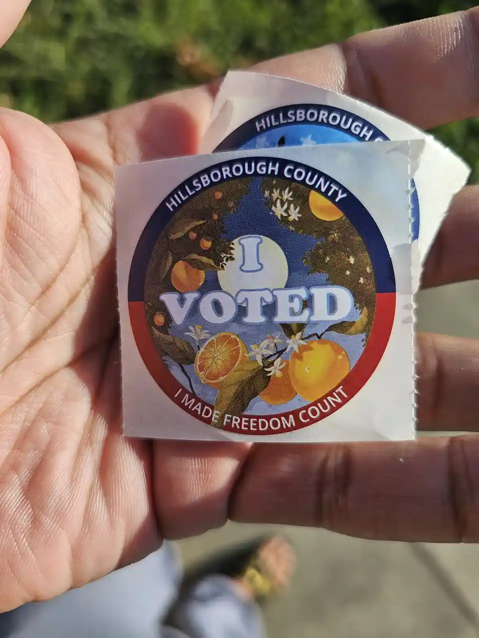 A hand holds a Hillsborough County 'I VOTED' sticker featuring oranges and citrus blossoms, with the slogan 'I MADE FREEDOM…
