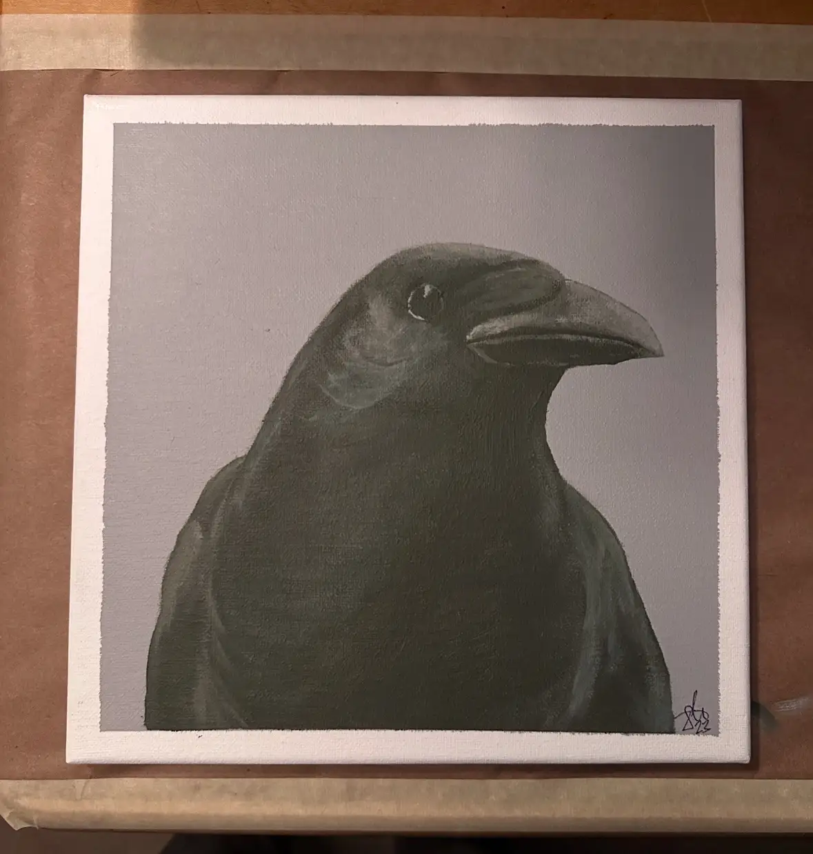 A grayscale painting of a crow's head and neck on a canvas, resting on brown paper. The crow is depicted in profile, looking…