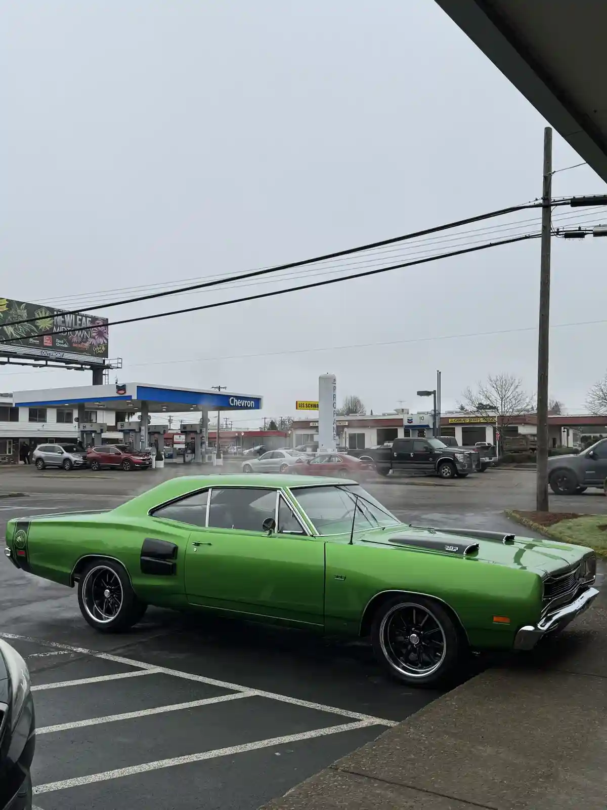 A bright green classic American muscle car, likely a Dodge Charger, is parked in a wet parking lot in front of a Chevron gas…