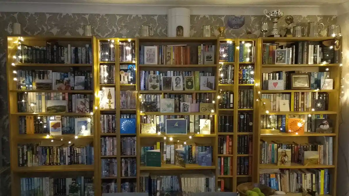 A cozy room features a large, multi-section wooden bookshelf filled with numerous books, decorative cards, and small…