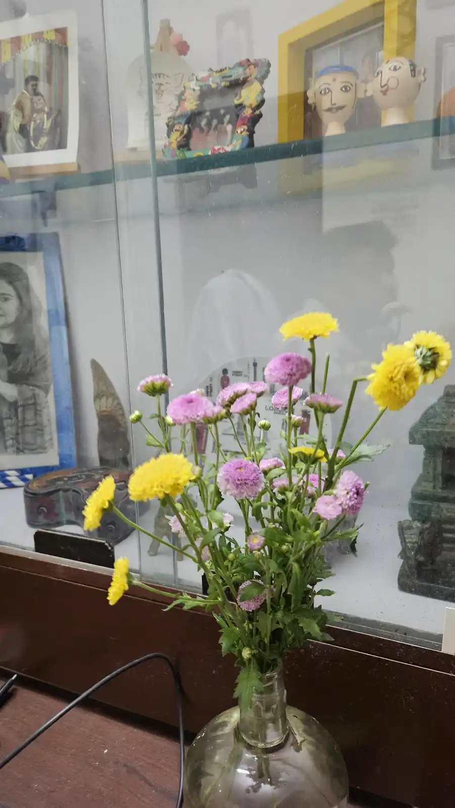 A vase filled with yellow and pink flowers sits on a wooden surface in front of a glass display case containing various…