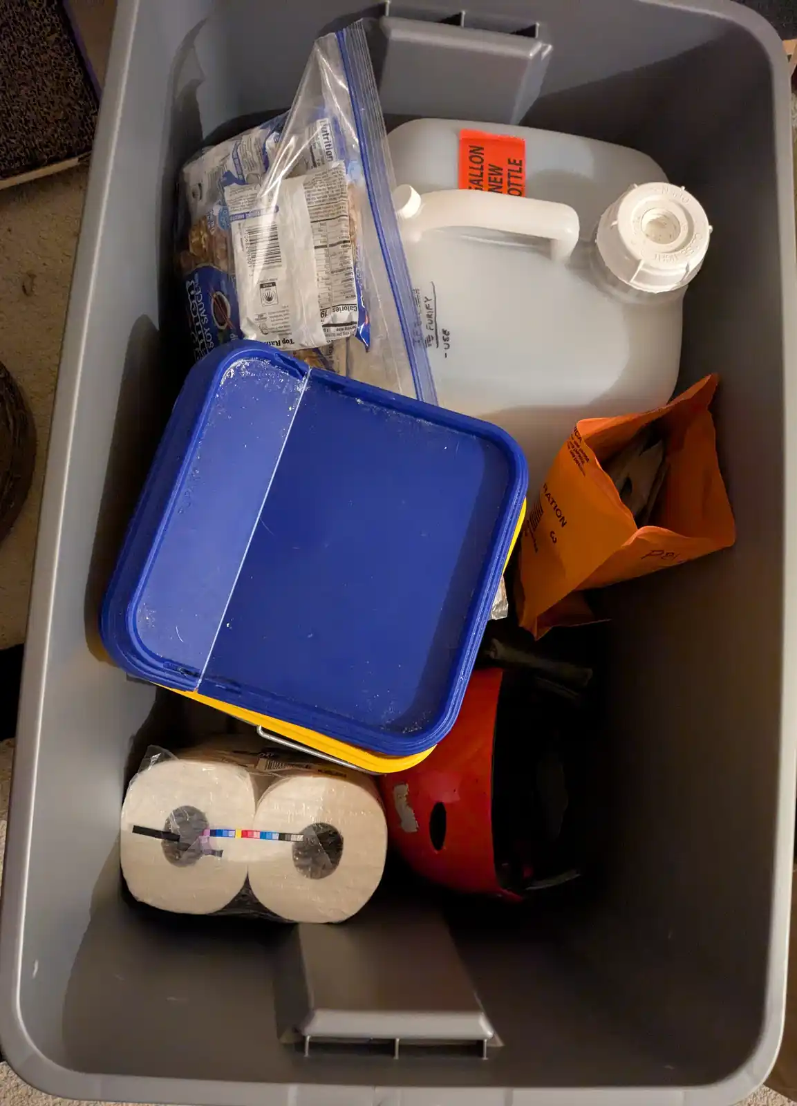 A gray plastic storage bin is filled with various household items including a large white water jug with an orange 'NEW…