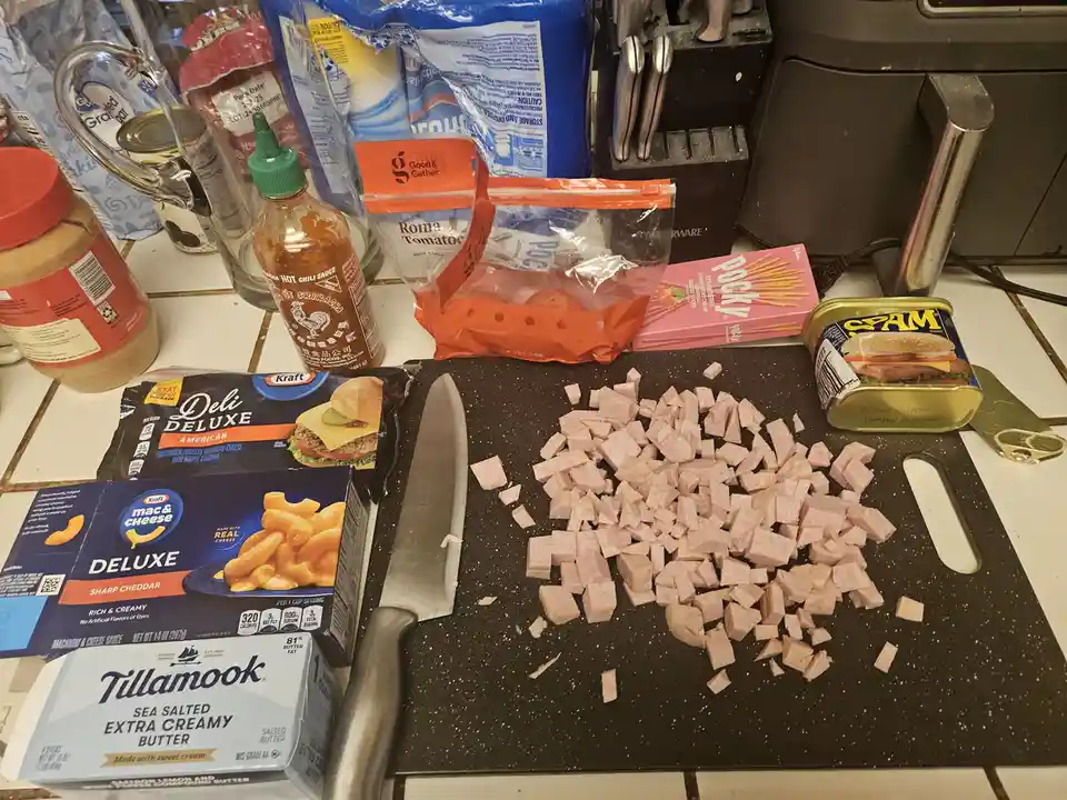 A cluttered kitchen counter displays a black cutting board with diced deli meat, a knife, and various food items including…