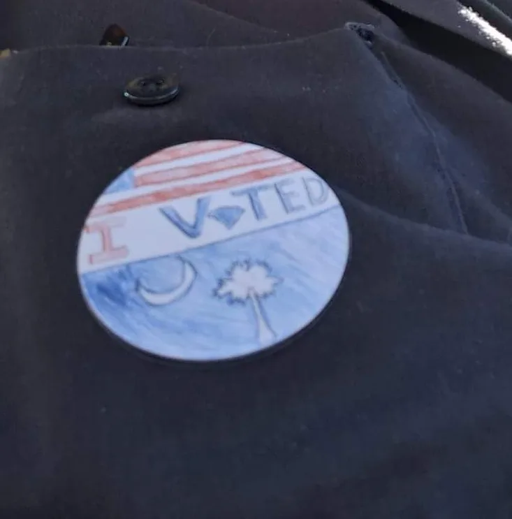 A close-up of a hand-drawn, circular 'I V-TED' button pinned to a dark jacket. The button features a red and white striped…