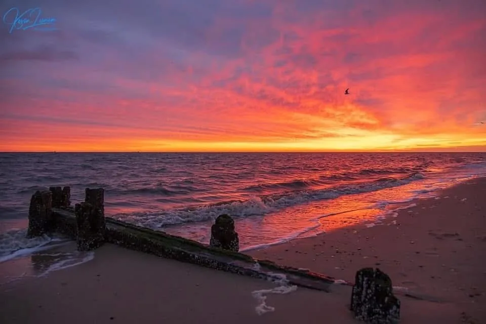 A serene beach sunset with vibrant orange and pink skies reflecting on the water, featuring weathered wooden pilings in the…