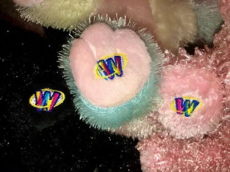 A close-up photo shows three fluffy, pink, heart-shaped plush objects, each with a colorful embroidered 'W' logo. One larger…