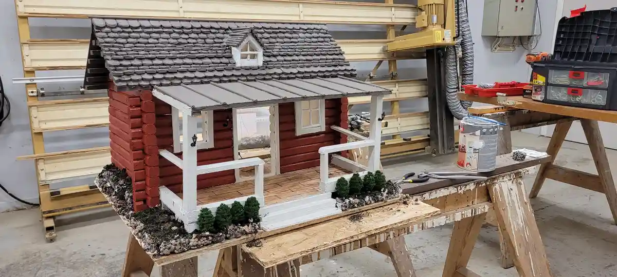 A meticulously crafted miniature red log cabin with a grey shingled roof and white porch sits on a wooden workbench in a…