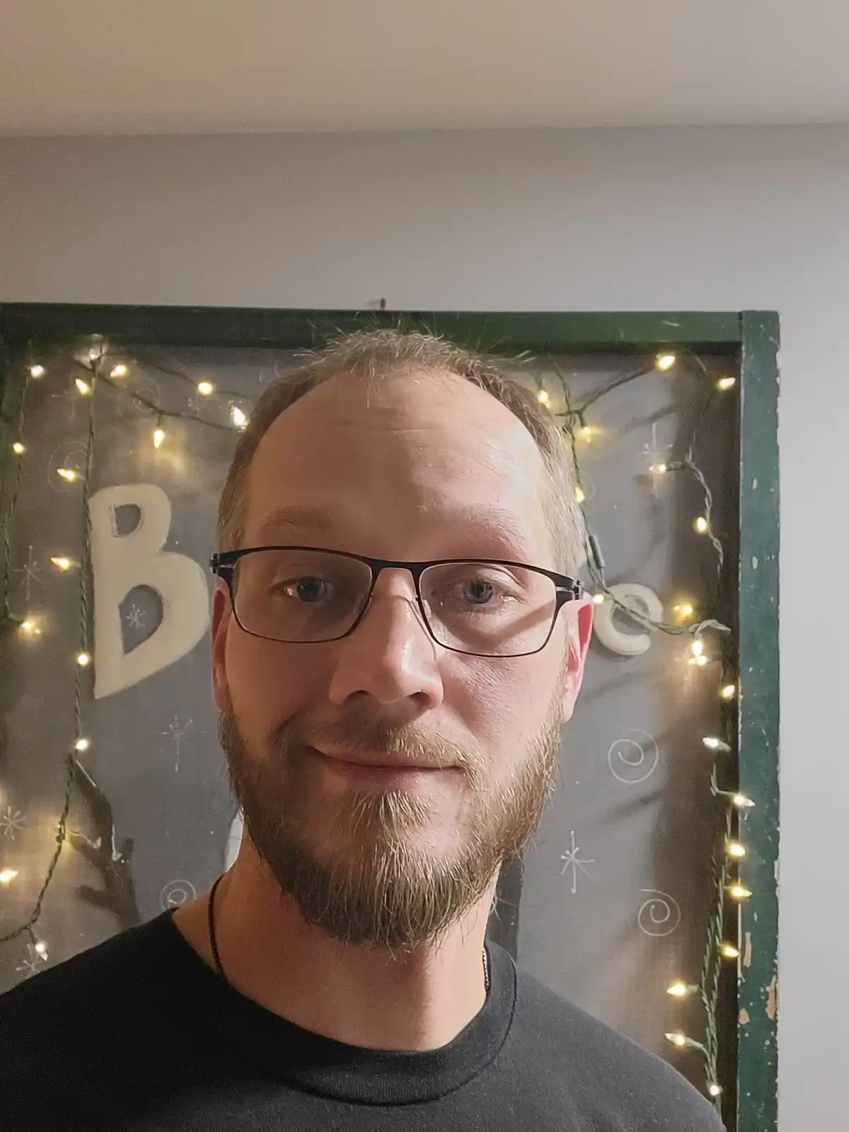 A man with glasses and a beard smiles at the camera, standing in front of a decorated green-framed window with string lights…