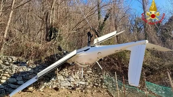 A white drone lies crashed on a rocky, wooded hillside, its wings broken and body damaged. The Italian Fire Department logo…