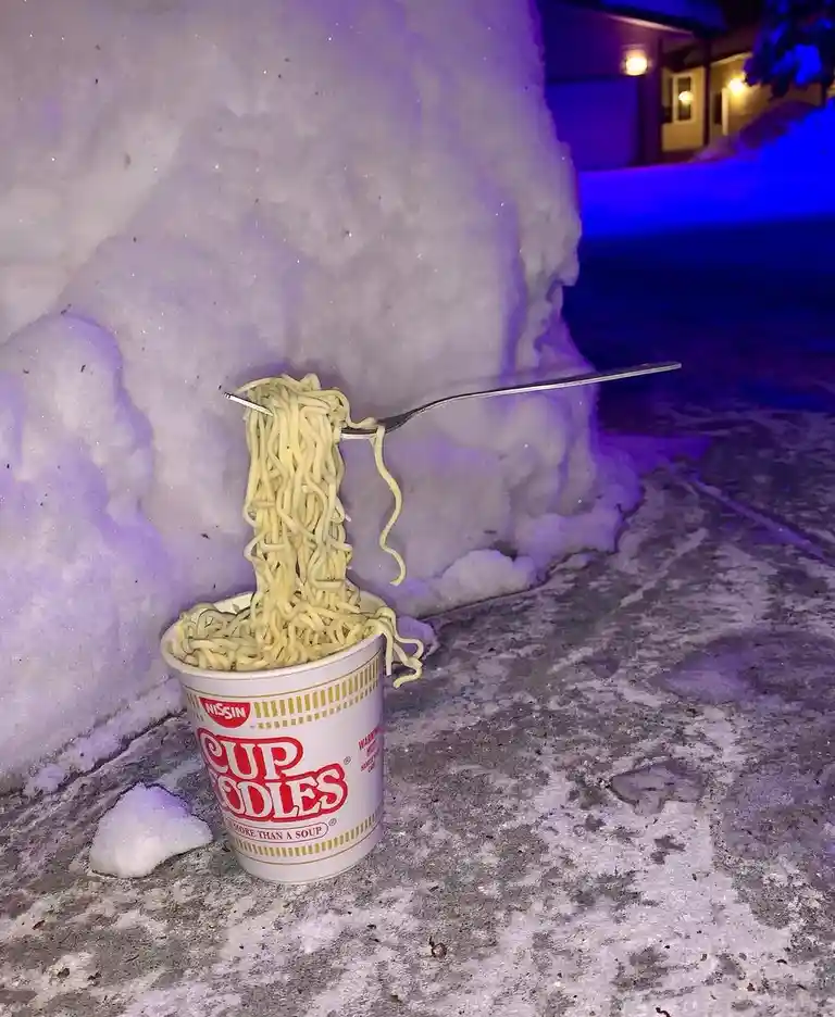 A cup of Nissin Cup Noodles sits on a snowy sidewalk at night, with a fork lifting noodles out of the cup. The background…