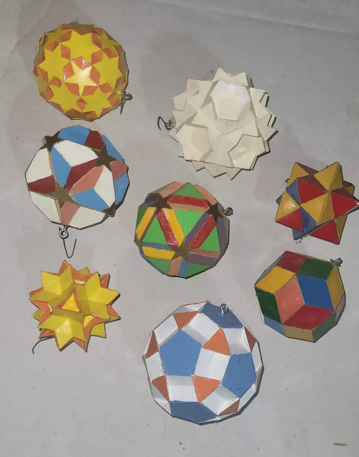 An overhead photo shows eight colorful, handcrafted geometric ornaments made of painted paper or cardboard, each with unique…