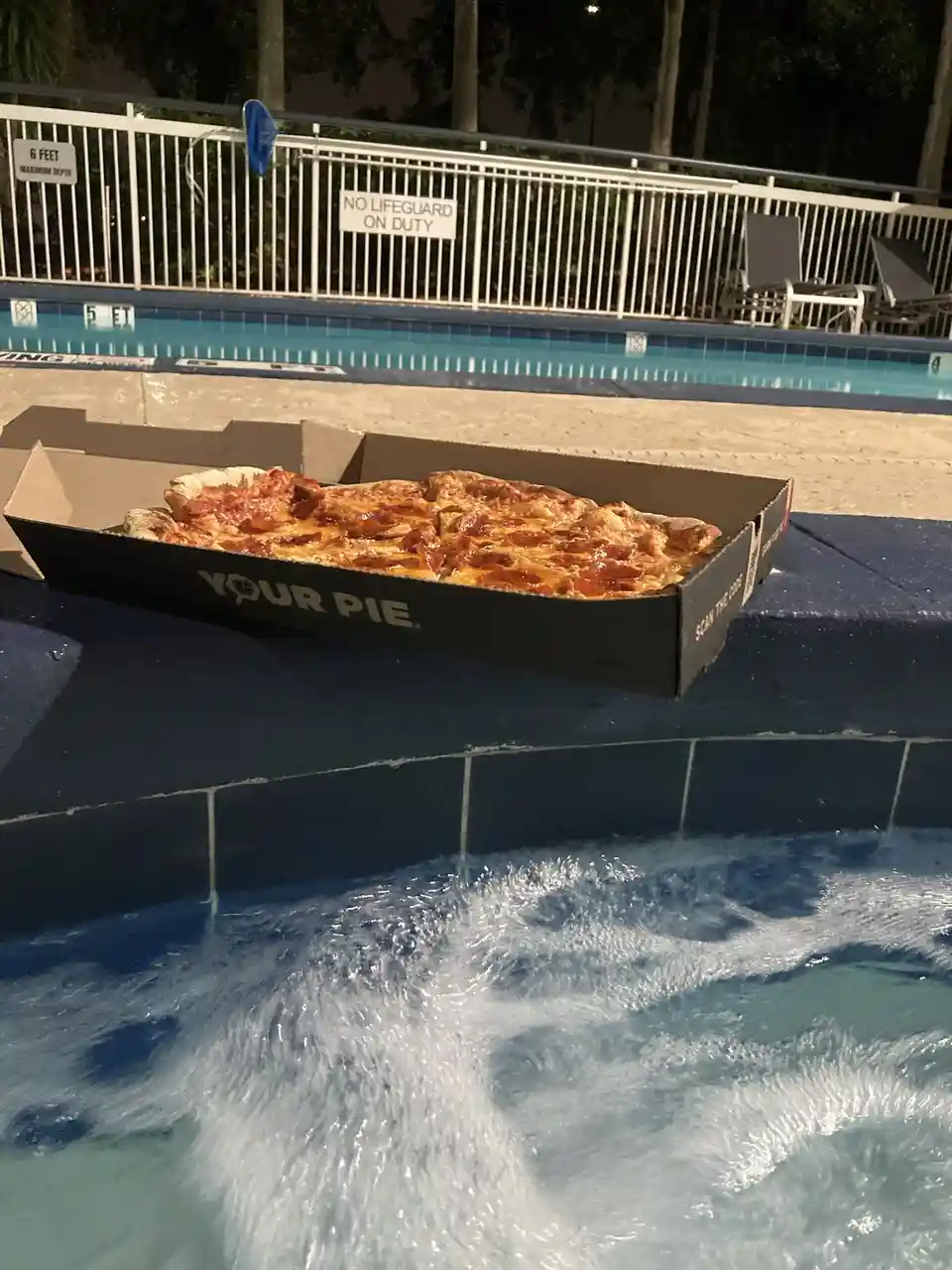 A pepperoni pizza in a black cardboard box labeled 'YOUR PIE' sits on the edge of a bubbling hot tub, with a swimming pool…