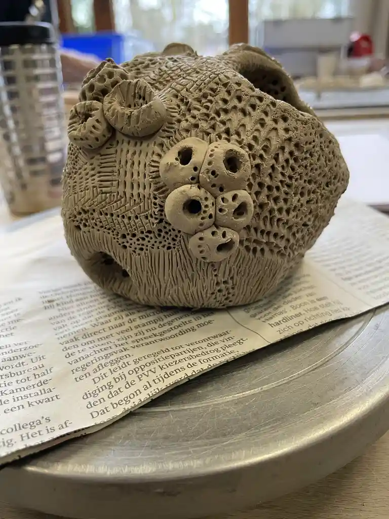 A close-up photo shows a textured, beige clay sculpture shaped like a bear's head, featuring detailed patterns, small holes…