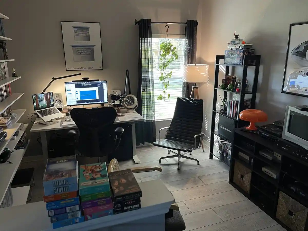 A well-lit home office features a desk with a computer setup, a black ergonomic chair, and a second, identical black chair…