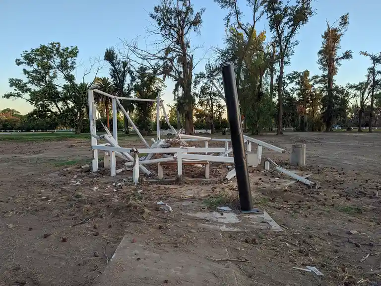 A photograph shows the wreckage of a white wooden structure, likely a swing set or play equipment, lying in disarray on dry…