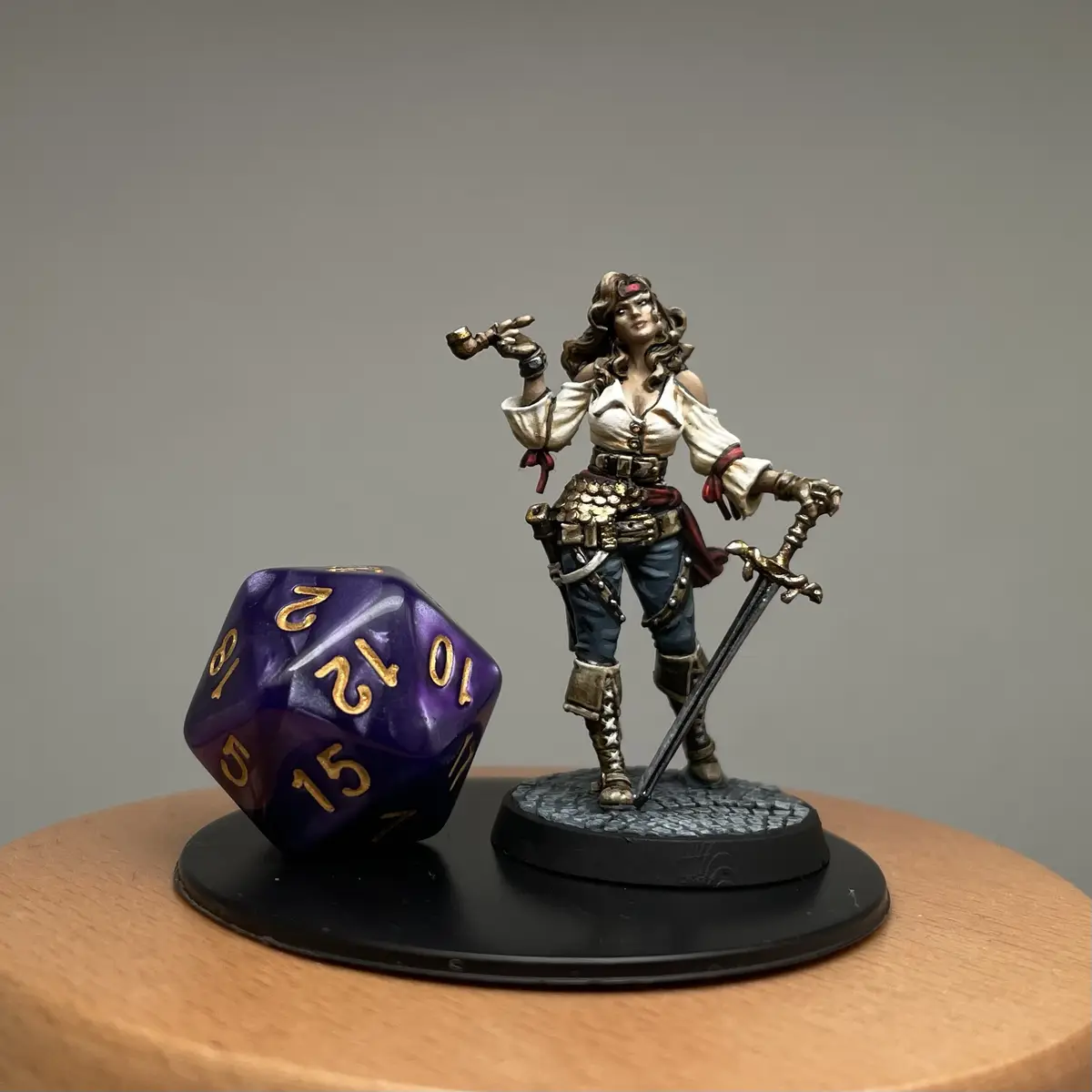 A painted miniature of a female pirate figure holding a sword and smoking a pipe stands next to a large purple 20-sided die…