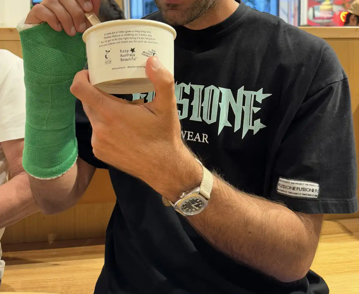A man with a green cast on his left arm holds a white ice cream cup with environmental messaging printed on the back. He…