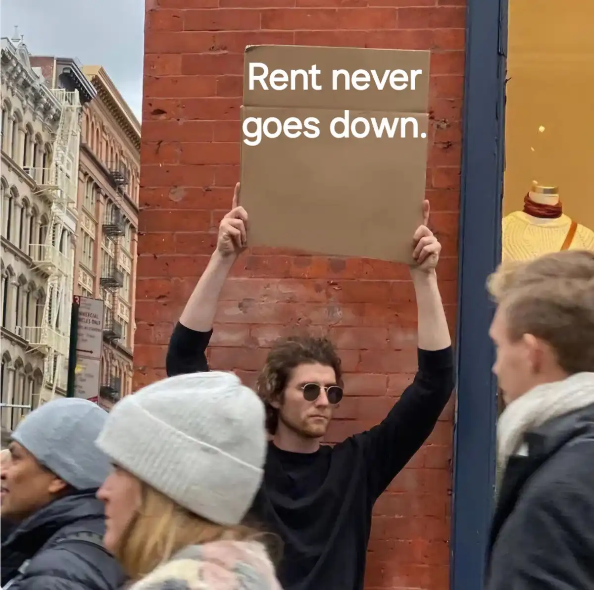 A person with curly hair and sunglasses holds up a cardboard sign that reads 'Rent never goes down.' in front of a red brick…