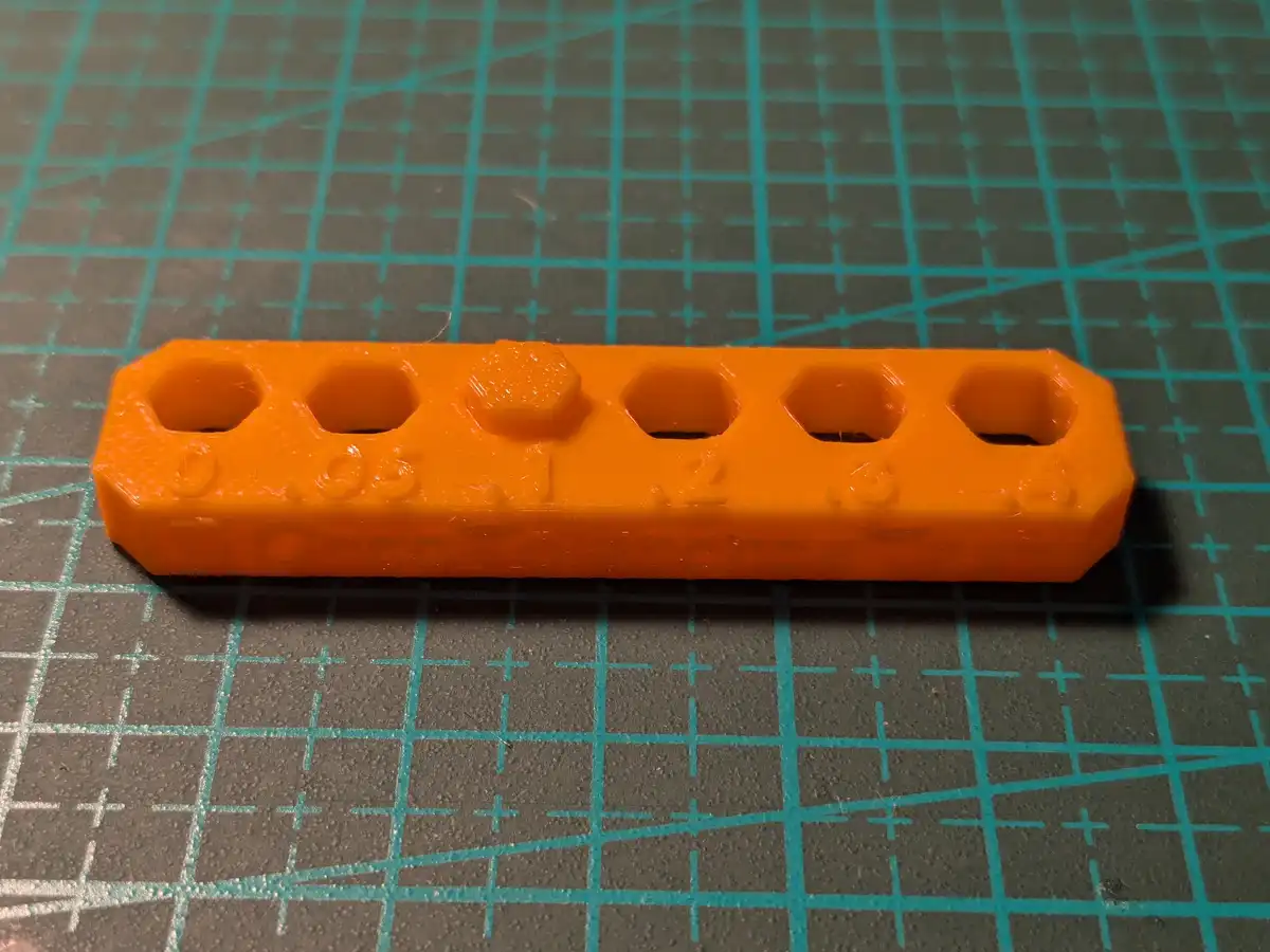 An orange 3D-printed plastic object with five hexagonal holes and embossed numbers 0, 0.5, 1, 2, and 3 along its length…