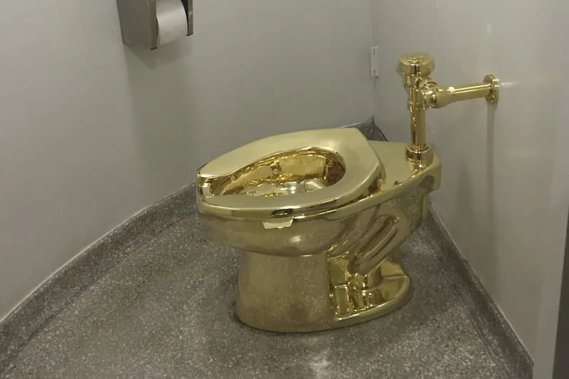 a toilet entirely coated in gold plating sits in a public restroom stall, with a standard toilet paper dispenser mounted on…