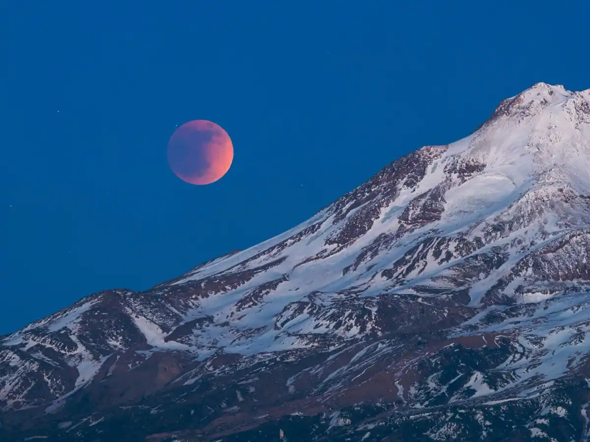 A dramatic night sky photo shows a reddish-orange moon, likely during a lunar eclipse, positioned above a snow-dusted…