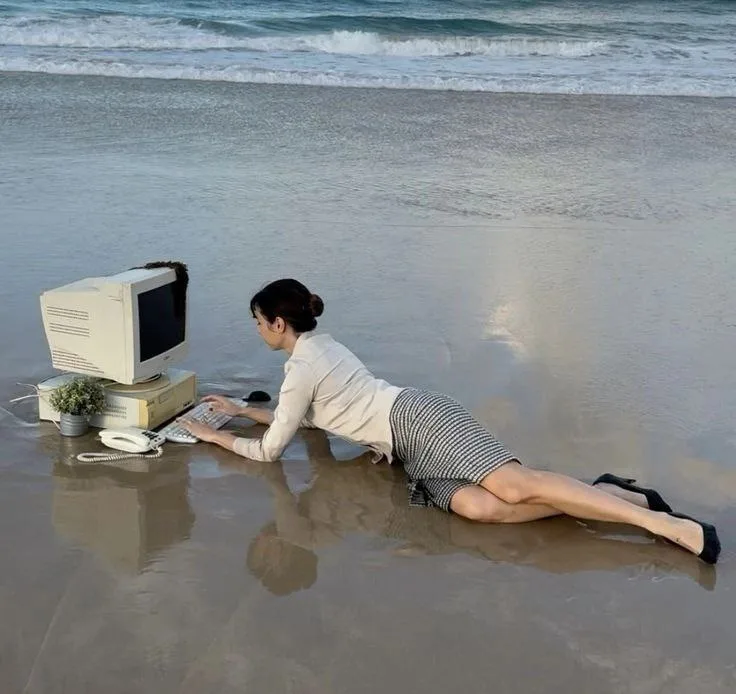A woman in business attire is lying prone on wet sand at the beach, typing on a vintage beige desktop computer with a CRT…