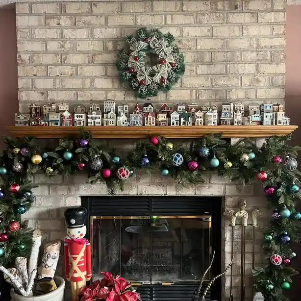 A fireplace mantel is heavily decorated for Christmas with a green wreath, colorful garland with ornaments, a nutcracker…
